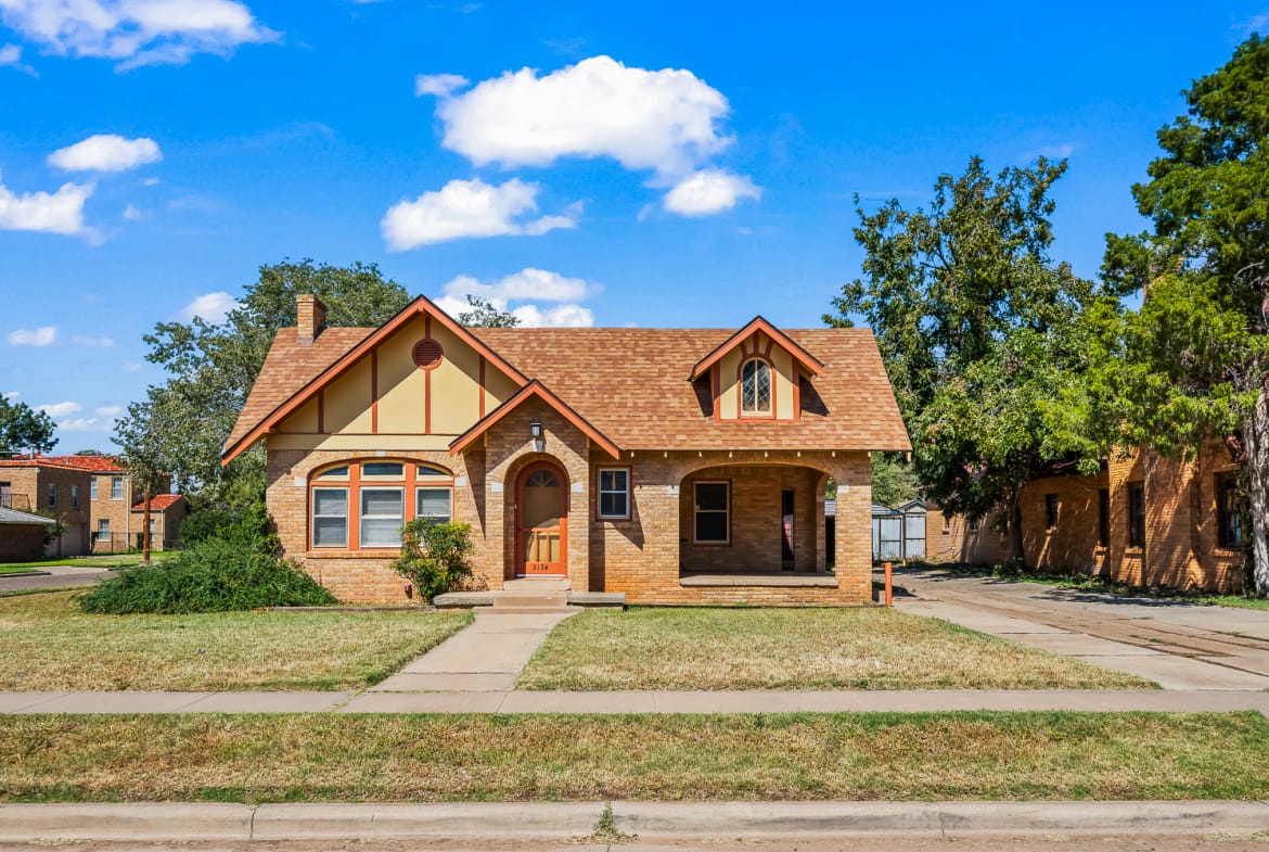Lubbock Lease Homes: A tan brick house with a steep, gabled roof, arched windows, and a small front porch sits under a bright blue sky. The yard has trimmed grass, a sidewalk, and some shrubs in front of the house.