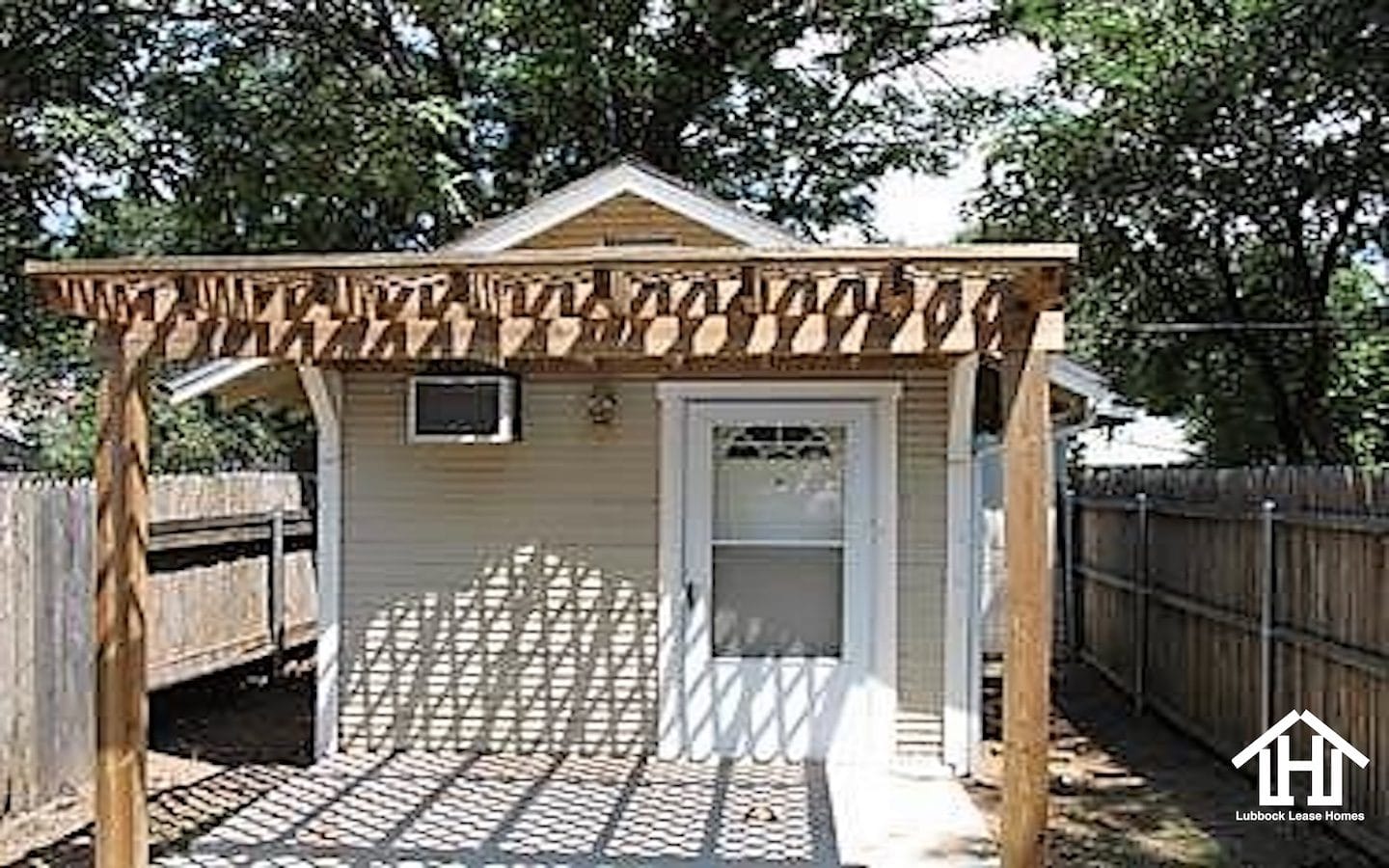 Lubbock Lease Homes: A small tan shed with a white door and window AC unit, topped by a wooden pergola casting shadows, is enclosed by a wooden fence and surrounded by trees.