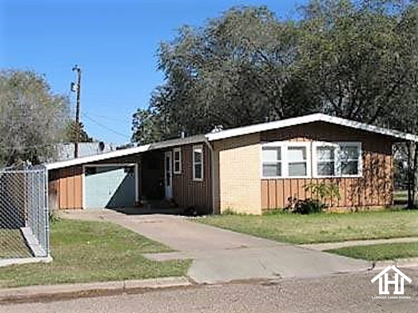 Lubbock Lease Homes: Single-story house with brown and beige siding, attached carport, driveway, green lawn, chain-link fence on the left, and trees in the background. A white logo is visible in the bottom right corner.