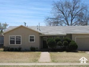 Lubbock Lease Homes: Single-story beige house with a gray roof, a small covered entry, trimmed bushes, and a large leafless tree in the background. The front yard has dry grass and a straight concrete walkway.
