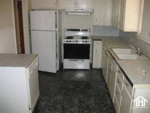 Lubbock Lease Homes: A small, old-fashioned kitchen with white cabinets, a white refrigerator, a gas stove, a dishwasher, and a double sink, all set against grayish countertops and dark patterned flooring.