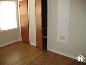 Lubbock Lease Homes: A small, empty room with light-colored walls, hardwood flooring, two wooden doors one open to a closet, a window with blinds, and a white utility panel on the wall.