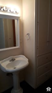 Lubbock Lease Homes: A small bathroom features a white pedestal sink with a mirror and lights above it, a towel ring on the wall, and built-in cabinets next to the sink.