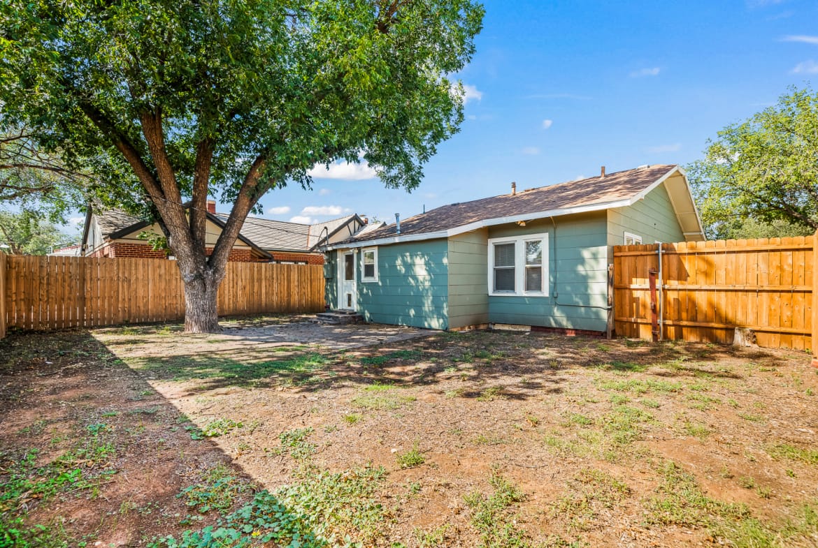 Lubbock Lease Homes: A fenced backyard with a large tree, patchy grass, and a light blue house with white trim and a small window. The yard is mostly bare soil with some green patches under a clear blue sky.