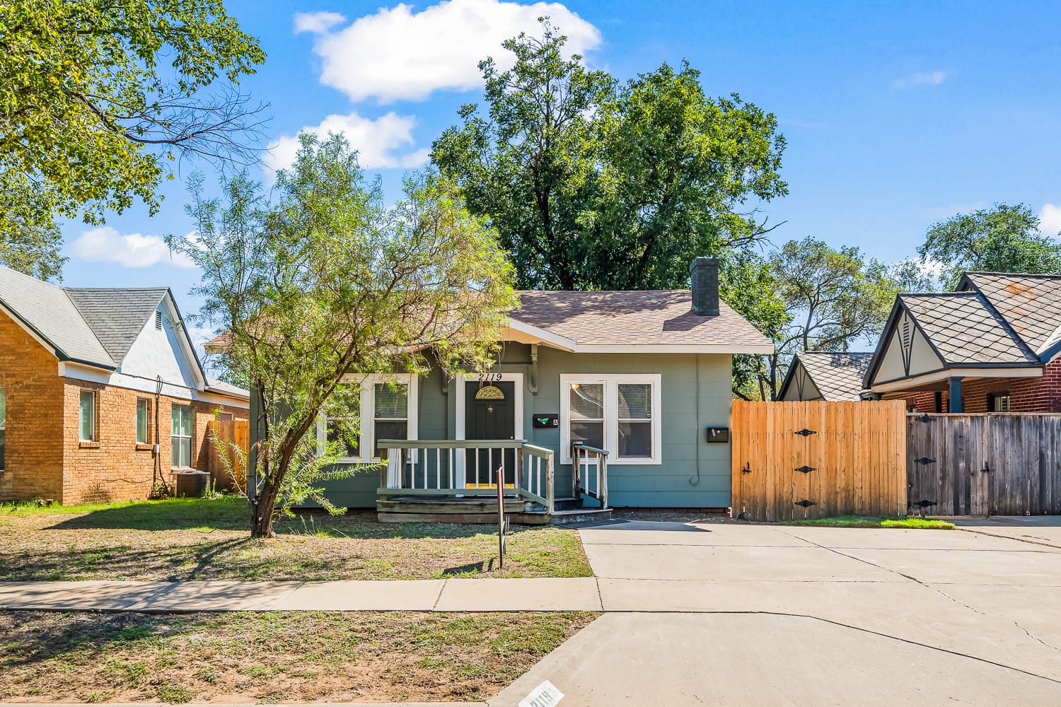 Lubbock Lease Homes: A small, light-blue house with a front porch, white trim, and a sloped roof sits between two neighboring homes. A tree and patchy lawn are in the front yard, and a driveway leads up to the porch.