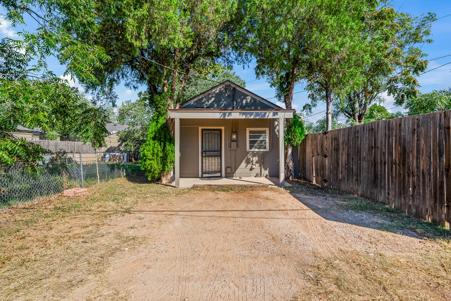 Lubbock Lease Homes: A small gray house with a covered front porch, metal screen door, and steps, surrounded by trees and a wooden fence, with a dirt driveway leading up to the entrance.