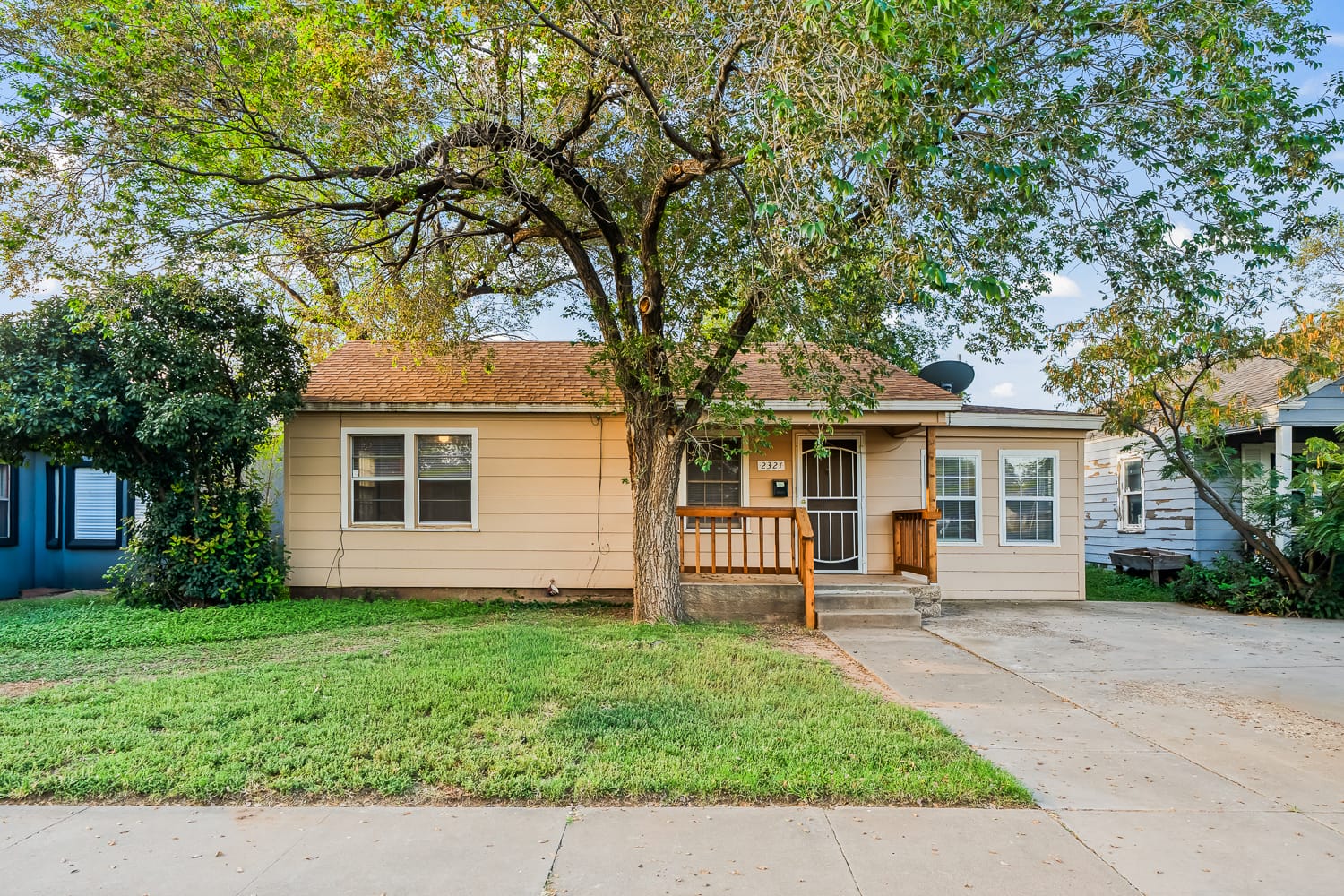 Lubbock Lease Homes: Single-story beige house with a gable roof, front porch, and wooden railings. A large tree shades the yard, and a driveway runs alongside the house. The lawn is green with a sidewalk in front.
