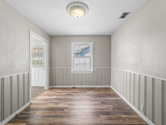 Lubbock Lease Homes: A small, empty room with beige walls, white trim, wood-look vinyl flooring, a single window, and a ceiling light. A door to the left leads outside, and the lower walls feature vertical paneling.