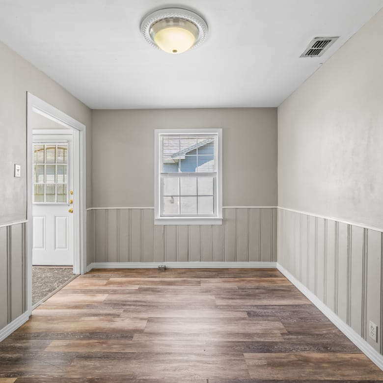 Lubbock Lease Homes: A small, empty room with beige walls, white trim, wood-look vinyl flooring, a single window, and a ceiling light. A door to the left leads outside, and the lower walls feature vertical paneling.