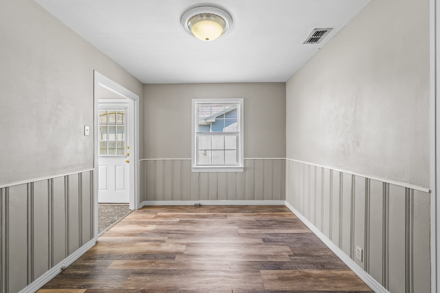 Lubbock Lease Homes: A small, empty room with beige walls, white trim, wood-look vinyl flooring, a single window, and a ceiling light. A door to the left leads outside, and the lower walls feature vertical paneling.