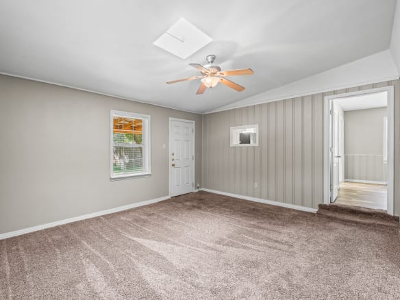 Lubbock Lease Homes: Spacious empty room with beige carpet, light gray walls, a white ceiling with a ceiling fan, a window, a white door, and an open doorway leading to another room with wood flooring.