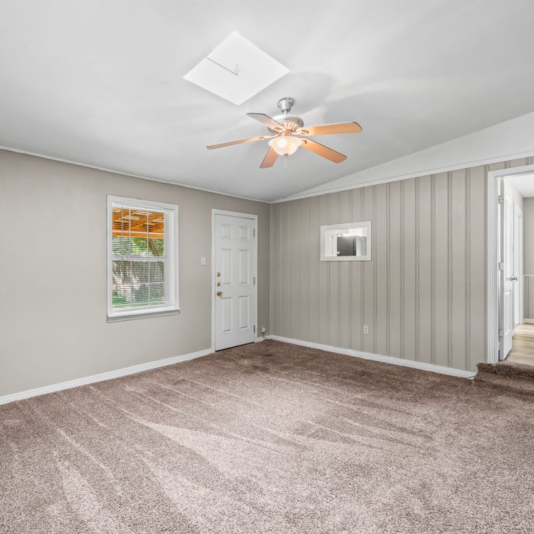 Lubbock Lease Homes: Spacious empty room with beige carpet, light gray walls, a white ceiling with a ceiling fan, a window, a white door, and an open doorway leading to another room with wood flooring.