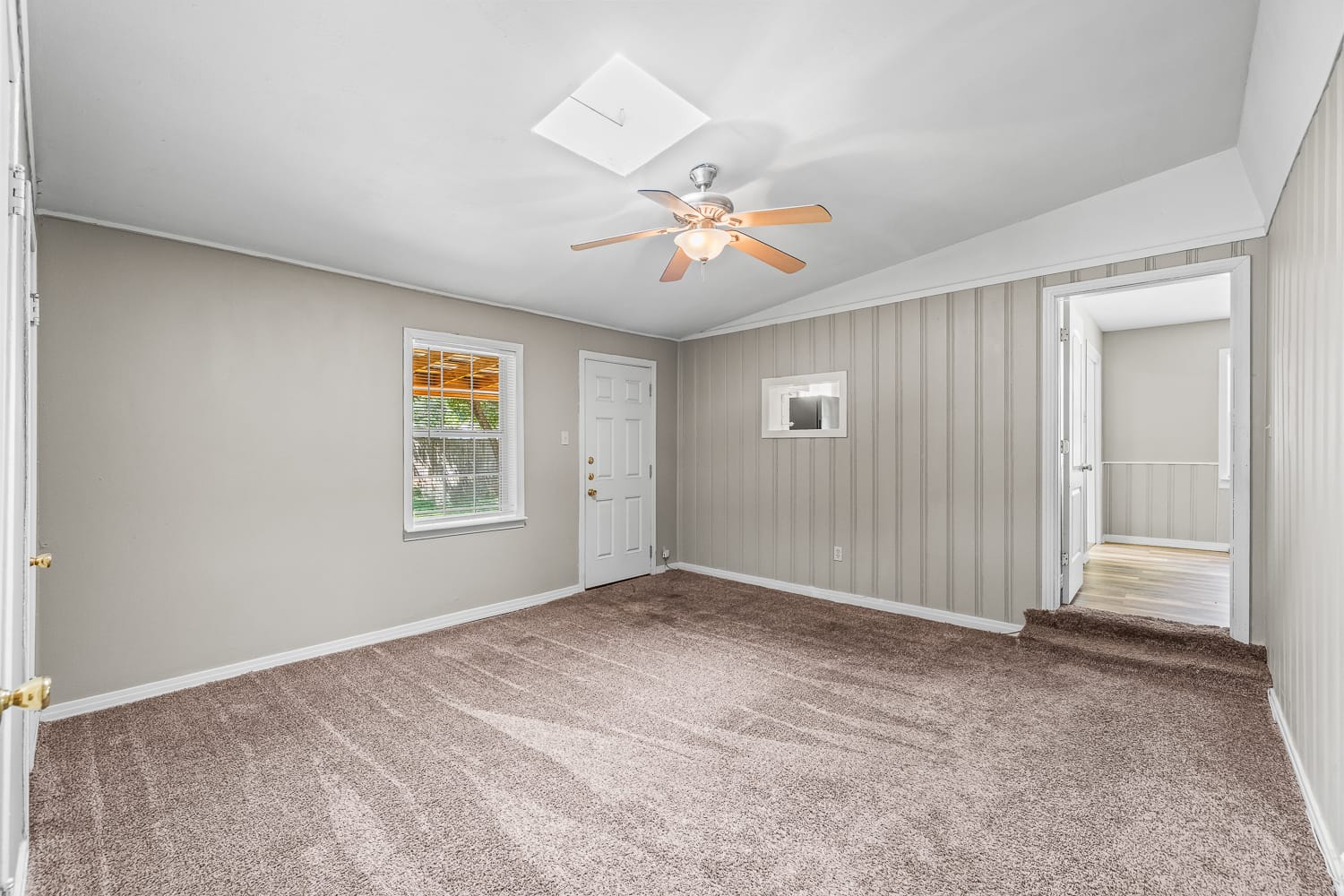 Lubbock Lease Homes: Spacious empty room with beige carpet, light gray walls, a white ceiling with a ceiling fan, a window, a white door, and an open doorway leading to another room with wood flooring.