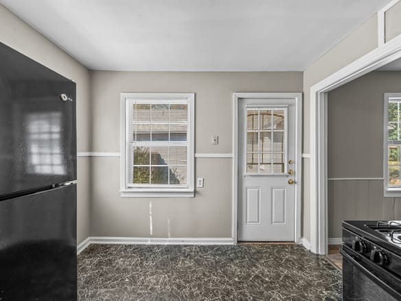 Lubbock Lease Homes: A small kitchen with a black refrigerator, a window with blinds, a white door with glass panels, beige walls, and a black and gray marbled floor. Part of a gas stove is visible on the right.