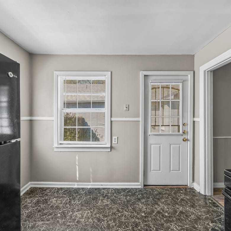 Lubbock Lease Homes: A small kitchen with a black refrigerator, a window with blinds, a white door with glass panels, beige walls, and a black and gray marbled floor. Part of a gas stove is visible on the right.