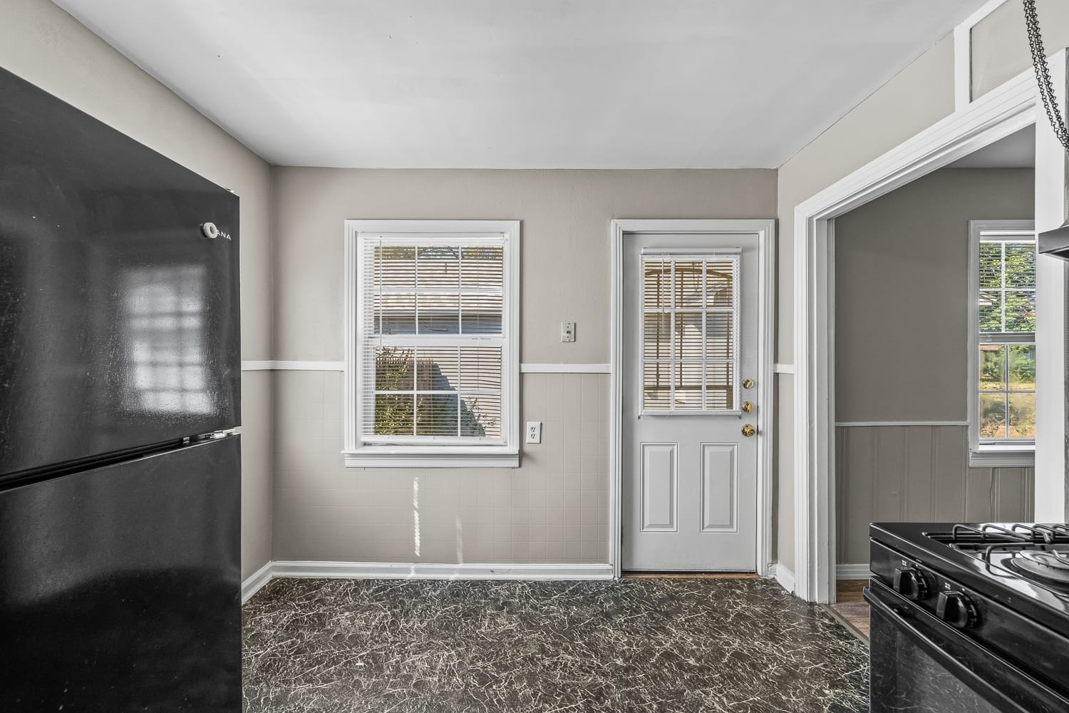 Lubbock Lease Homes: A small kitchen with a black refrigerator, a window with blinds, a white door with glass panels, beige walls, and a black and gray marbled floor. Part of a gas stove is visible on the right.