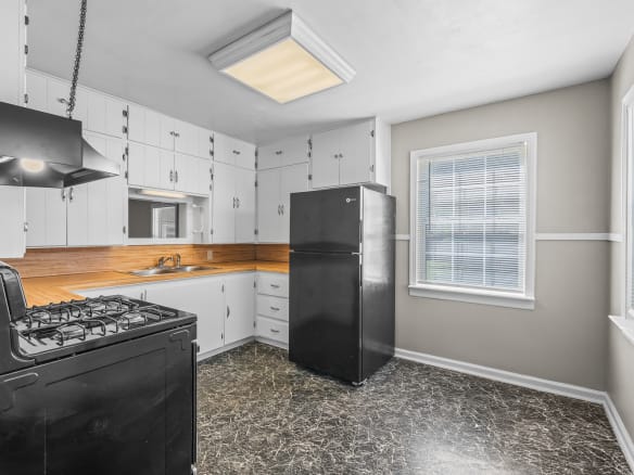 Lubbock Lease Homes: Bright kitchen with white cabinets, wood countertop, black stove, and black refrigerator. Large window lets in natural light, and the floor is dark with a marbled pattern. Walls are light with white trim.