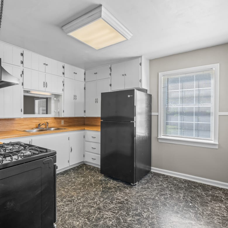 Lubbock Lease Homes: Bright kitchen with white cabinets, wood countertop, black stove, and black refrigerator. Large window lets in natural light, and the floor is dark with a marbled pattern. Walls are light with white trim.