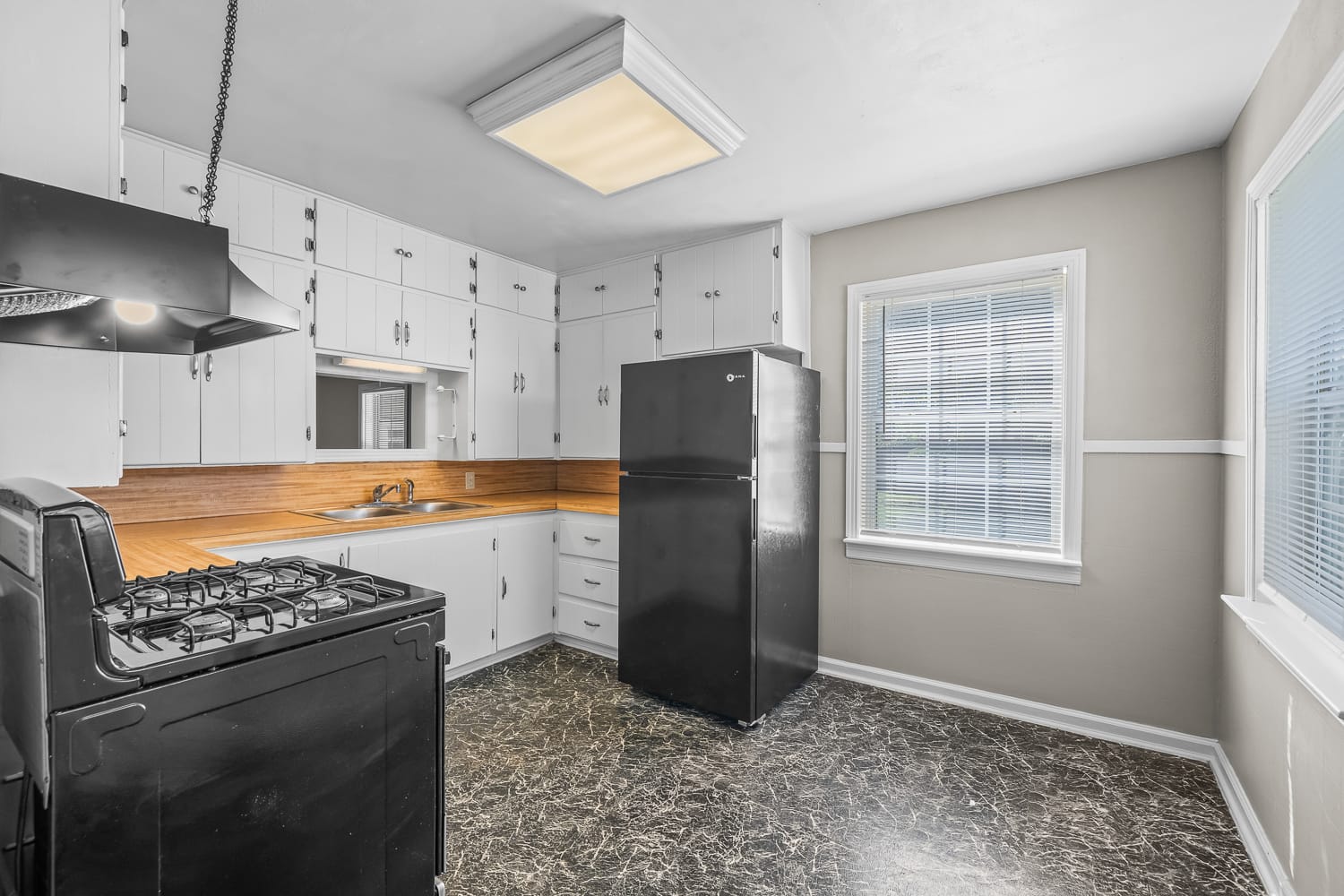 Lubbock Lease Homes: Bright kitchen with white cabinets, wood countertop, black stove, and black refrigerator. Large window lets in natural light, and the floor is dark with a marbled pattern. Walls are light with white trim.