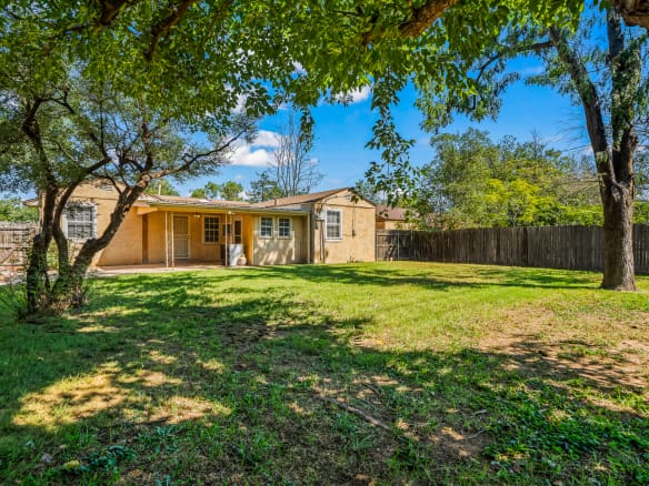 Lubbock Lease Homes: A spacious backyard with green grass, scattered shade from large trees, and a wooden fence surrounding a single-story yellow house with a covered patio.