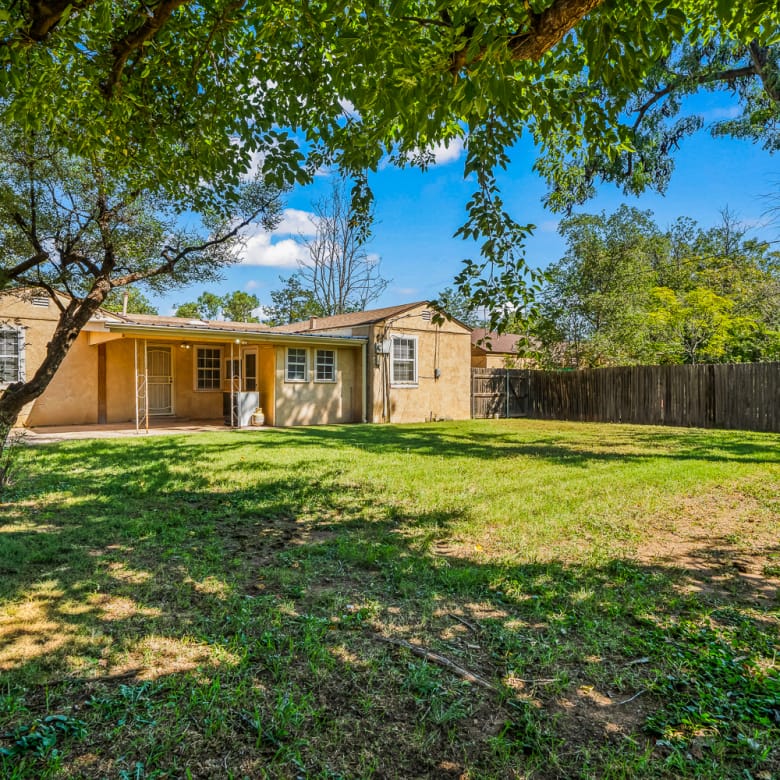 Lubbock Lease Homes: A spacious backyard with green grass, scattered shade from large trees, and a wooden fence surrounding a single-story yellow house with a covered patio.