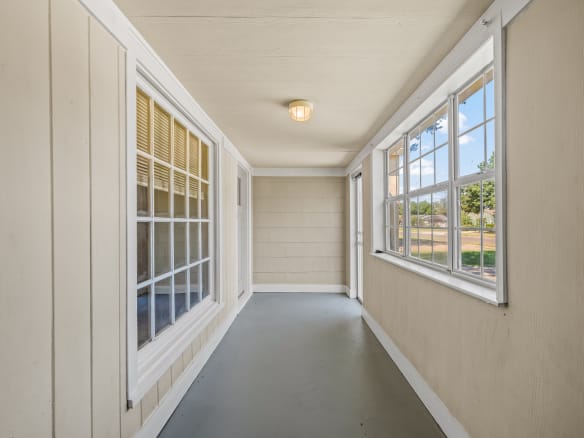 Lubbock Lease Homes: A narrow, enclosed porch with beige paneled walls, large windows on both sides, a gray floor, and a ceiling light. Sunlight streams in through the windows, creating a bright and airy space.