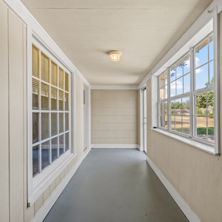 Lubbock Lease Homes: A narrow, enclosed porch with beige paneled walls, large windows on both sides, a gray floor, and a ceiling light. Sunlight streams in through the windows, creating a bright and airy space.