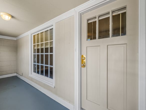 Lubbock Lease Homes: A beige door with a gold handle and small square windows sits beside a large white-trimmed window on a covered porch with light tan paneled walls and a gray floor.
