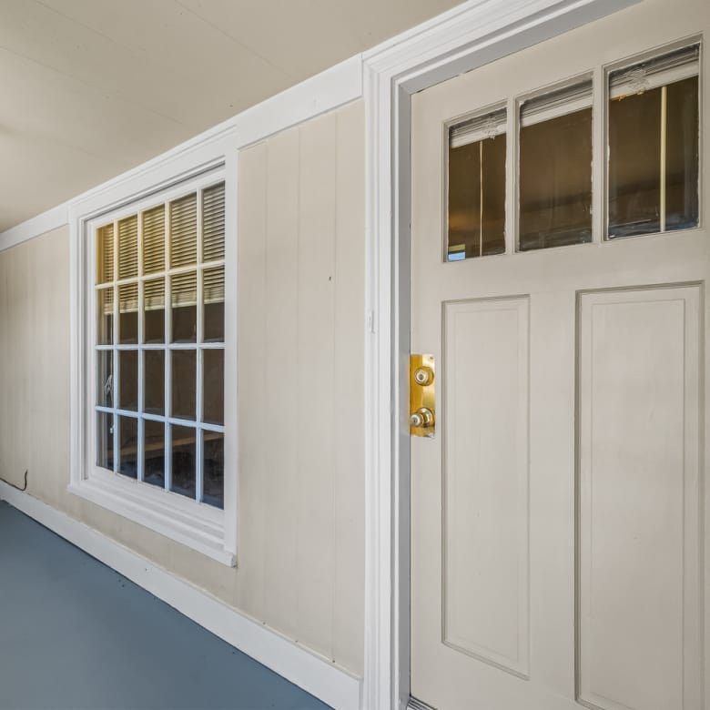 Lubbock Lease Homes: A beige door with a gold handle and small square windows sits beside a large white-trimmed window on a covered porch with light tan paneled walls and a gray floor.