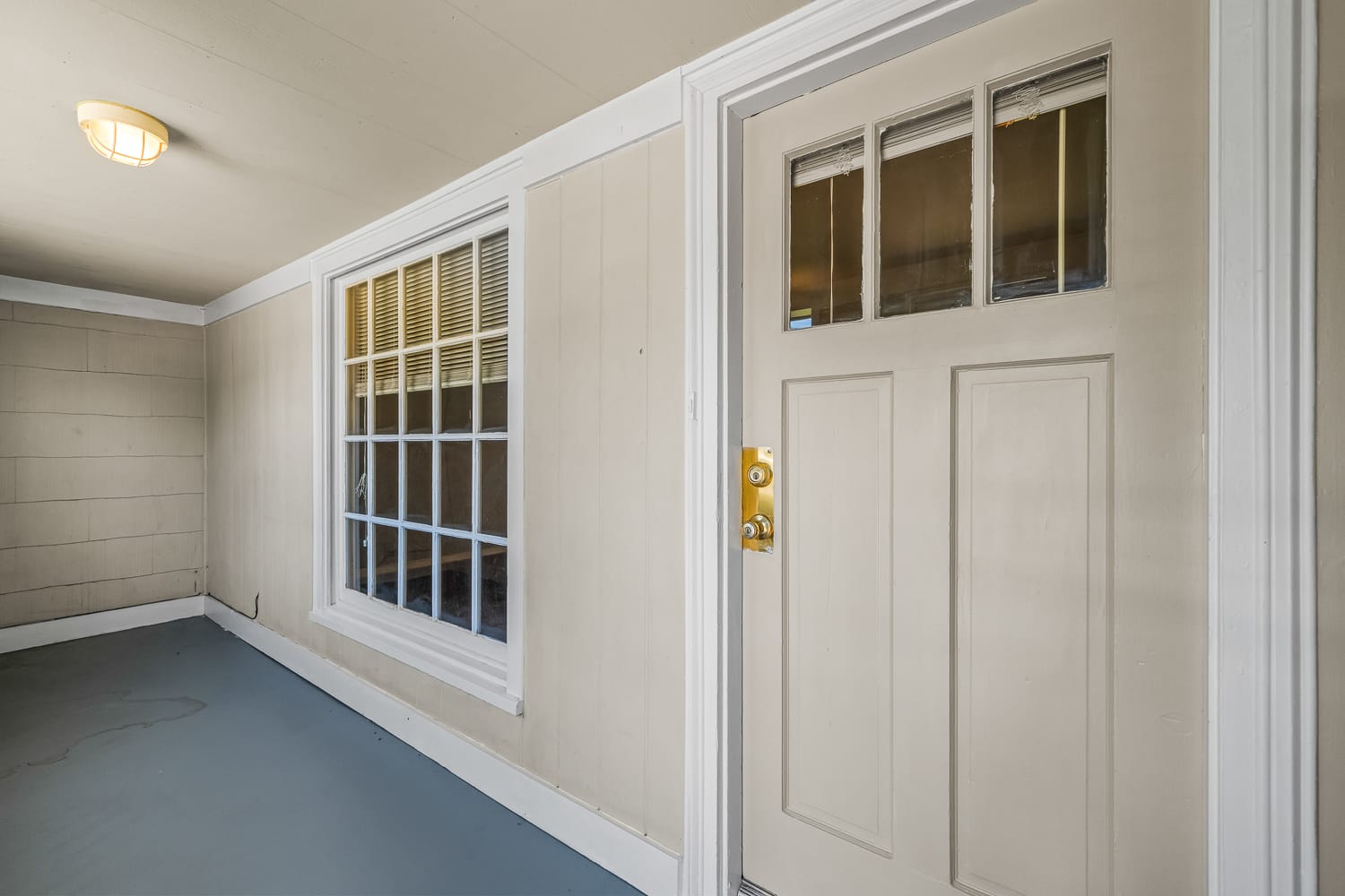 Lubbock Lease Homes: A beige door with a gold handle and small square windows sits beside a large white-trimmed window on a covered porch with light tan paneled walls and a gray floor.
