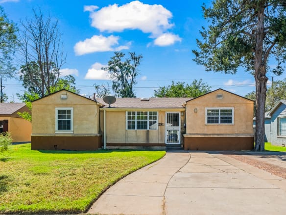Lubbock Lease Homes: A small, single-story beige house with a split facade, front lawn, gated entrance, and curved driveway under a bright blue sky with scattered clouds.