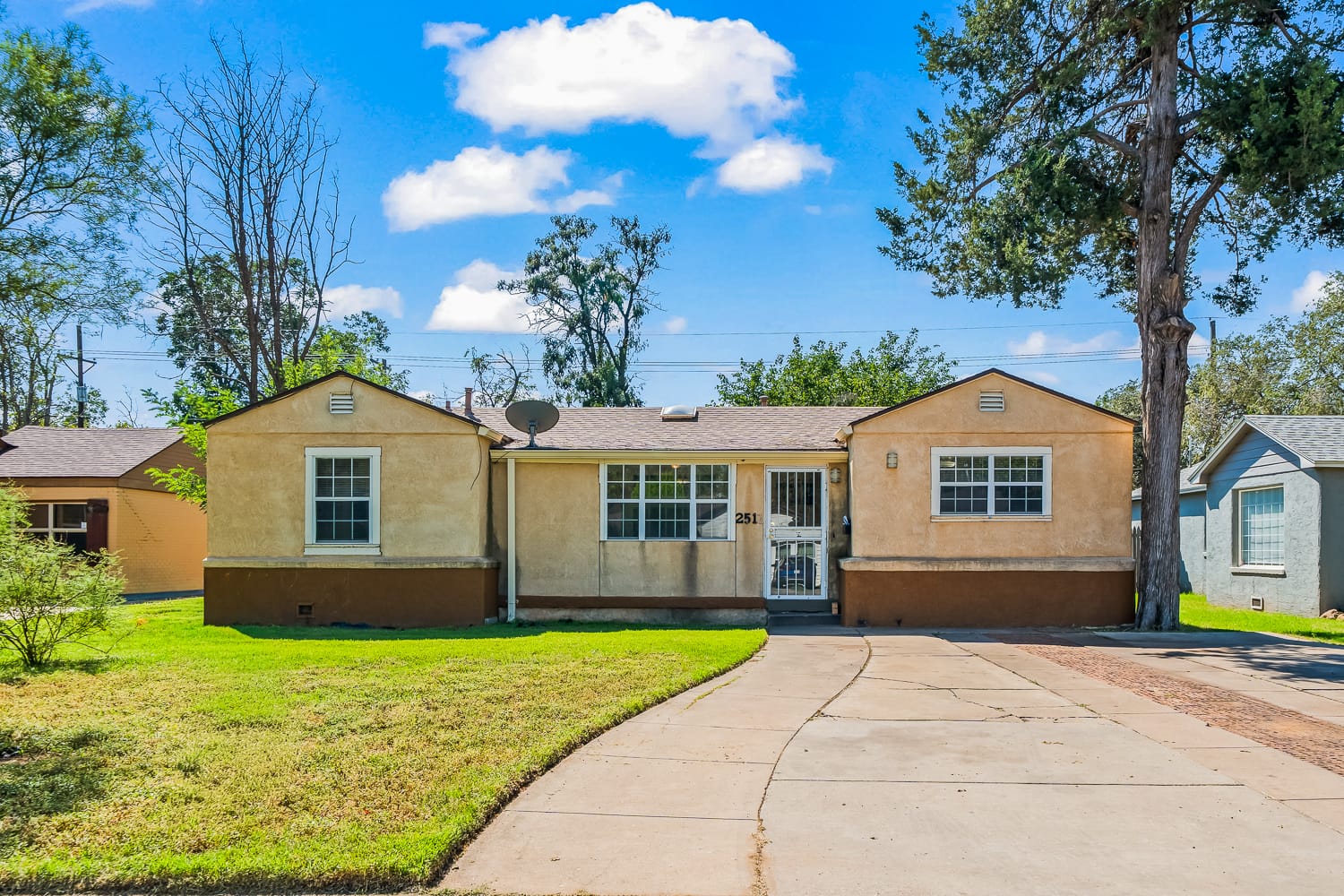 Lubbock Lease Homes: A small, single-story beige house with a split facade, front lawn, gated entrance, and curved driveway under a bright blue sky with scattered clouds.