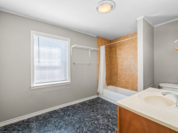 Lubbock Lease Homes: A bathroom with gray walls, a window with blinds, a white bathtub and shower with a beige tile wall, a white toilet, a single sink vanity, and black marbled flooring. A towel rack is mounted near the window.