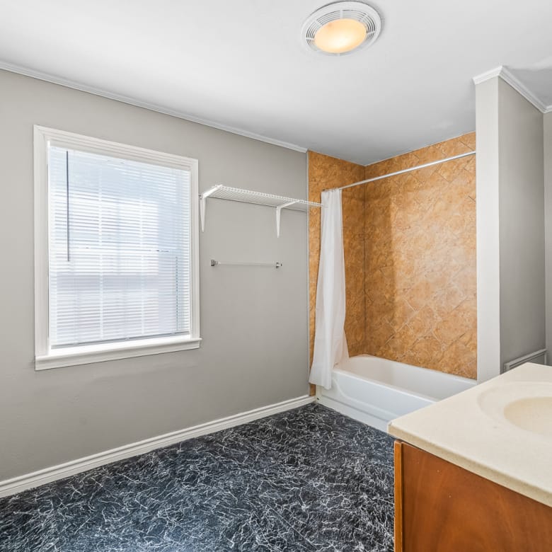 Lubbock Lease Homes: A bathroom with gray walls, a window with blinds, a white bathtub and shower with a beige tile wall, a white toilet, a single sink vanity, and black marbled flooring. A towel rack is mounted near the window.