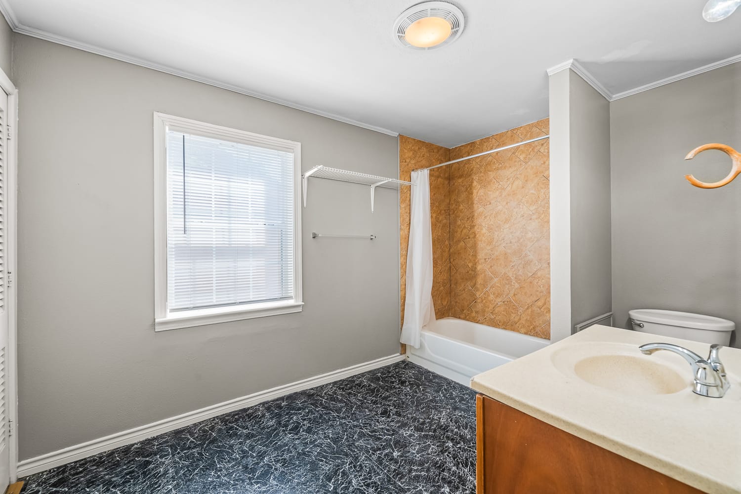 Lubbock Lease Homes: A bathroom with gray walls, a window with blinds, a white bathtub and shower with a beige tile wall, a white toilet, a single sink vanity, and black marbled flooring. A towel rack is mounted near the window.