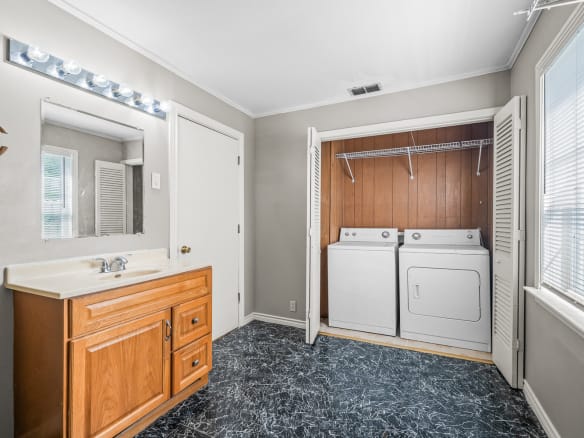 Lubbock Lease Homes: A laundry room with a wood cabinet sink, a mirror above with vanity lights, and a washer and dryer set in a closet with louvered doors. The floor has a black and white marble pattern. Sunlight streams through windows.