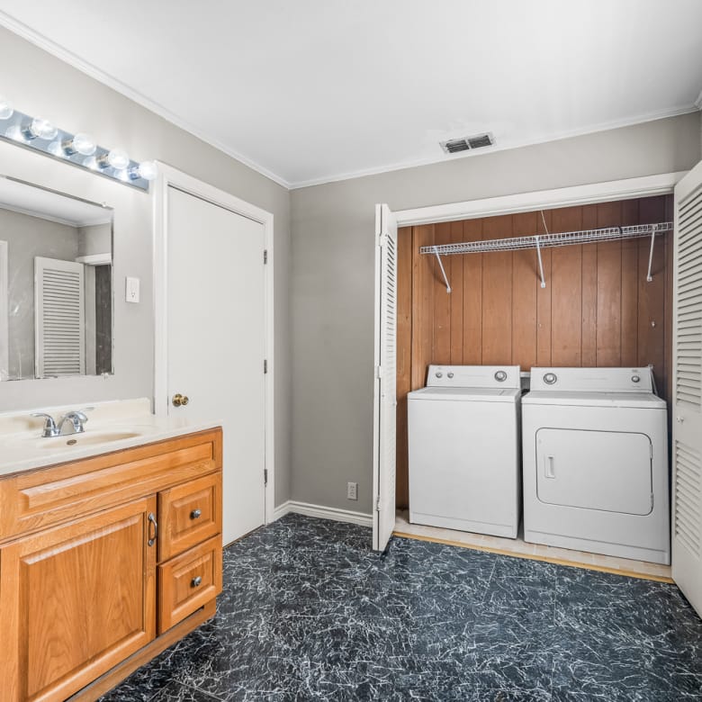 Lubbock Lease Homes: A laundry room with a wood cabinet sink, a mirror above with vanity lights, and a washer and dryer set in a closet with louvered doors. The floor has a black and white marble pattern. Sunlight streams through windows.