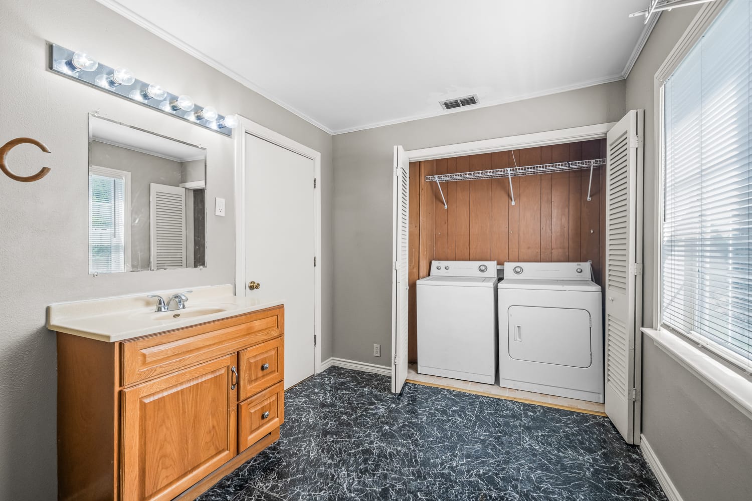 Lubbock Lease Homes: A laundry room with a wood cabinet sink, a mirror above with vanity lights, and a washer and dryer set in a closet with louvered doors. The floor has a black and white marble pattern. Sunlight streams through windows.