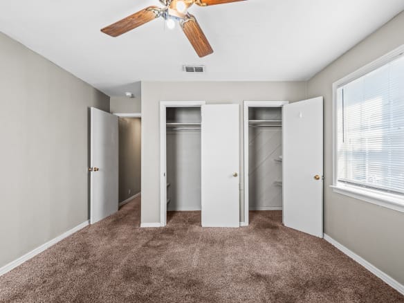 Lubbock Lease Homes: A bedroom with beige walls, brown carpet, a ceiling fan with lights, two open closets, an open door to a hallway, and a window letting in natural light.