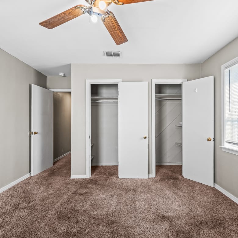 Lubbock Lease Homes: A bedroom with beige walls, brown carpet, a ceiling fan with lights, two open closets, an open door to a hallway, and a window letting in natural light.
