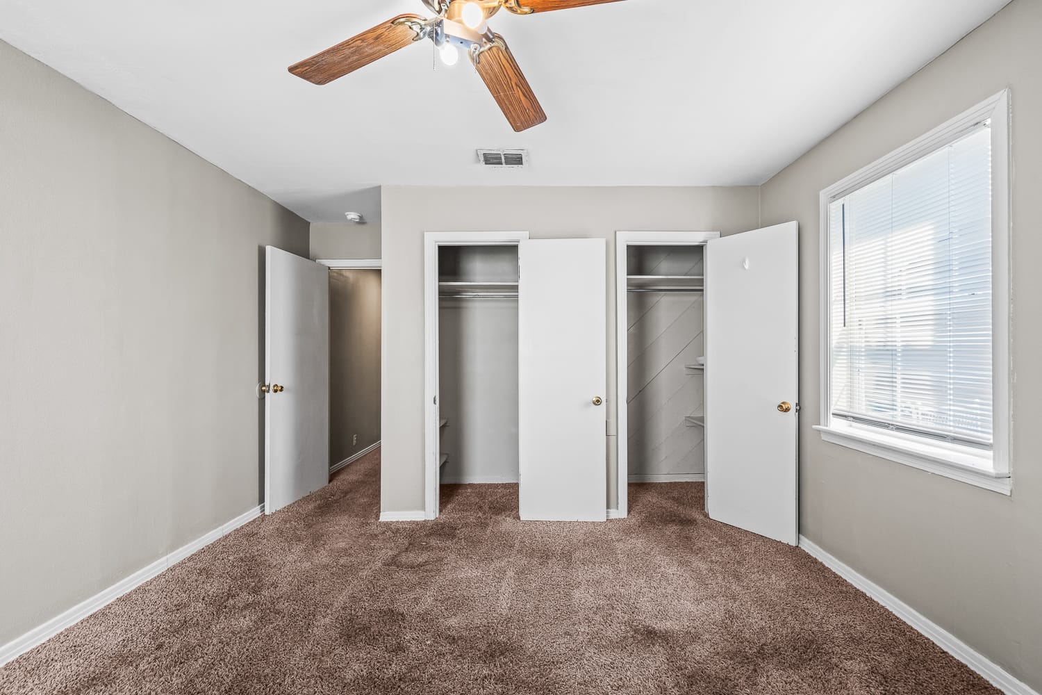 Lubbock Lease Homes: A bedroom with beige walls, brown carpet, a ceiling fan with lights, two open closets, an open door to a hallway, and a window letting in natural light.