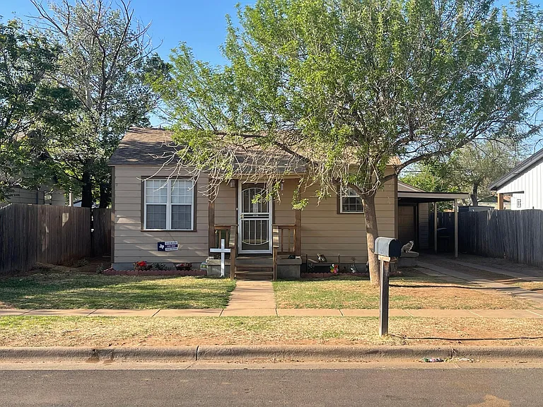 Lubbock Lease Homes: A small, tan house with a front porch, two steps, and a white front door. There are a few trees and flower beds in the yard, a mailbox by the curb, and a “For Sale” sign near the walkway.