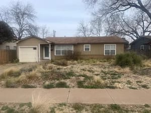 Lubbock Lease Homes: A single-story house with a brown exterior, attached garage, and a large, overgrown front yard featuring dry grass and shrubs. Leafless trees are visible in the background under a cloudy sky.