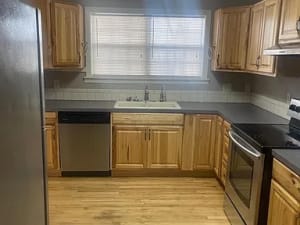 Lubbock Lease Homes: A kitchen with light wood cabinets, stainless steel appliances, a double sink under a window with blinds, black countertops, and a light wood floor.