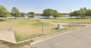 Lubbock Lease Homes: A paved path curves through a grassy park next to a calm lake, with trees and buildings in the background under a clear blue sky. A handicapped parking sign stands near the sidewalk—a peaceful scene for anyone moving to Lubbock.