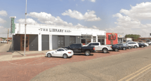 Lubbock Lease Homes: A row of parked cars lines a street in front of The Library Bar, a white brick building with large windows. The sky is partly cloudy, and a red sign reading LIT is visible next door—classic downtown charm amid growing Lubbock safety efforts.