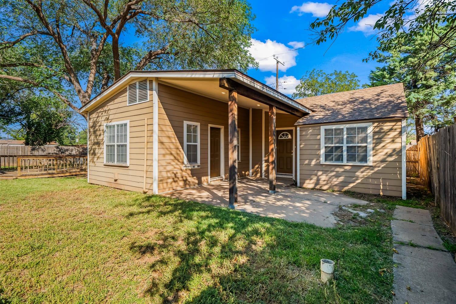 Lubbock Lease Homes: Small tan house with a covered patio, white trim, and large windows, surrounded by a grassy yard and enclosed by a wooden fence and trees under a blue sky with scattered clouds.