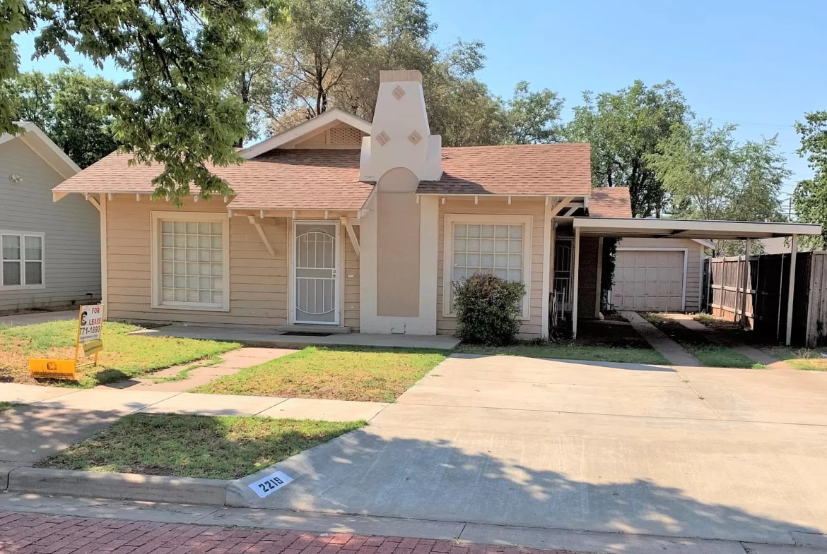 Lubbock Lease Homes: Single-story beige house with a brown roof, white trim, and decorative chimney. There’s a front lawn, a small bush, a driveway, a carport, and a garage in the background. A For Sale sign is posted on the lawn.