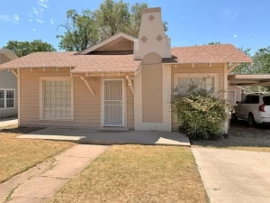 Lubbock Lease Homes: A small beige house with a brown roof, decorative chimney, white-trimmed windows, and a white door. A car is parked in the driveway on the right, and the lawn is dry and patchy under a clear sky.