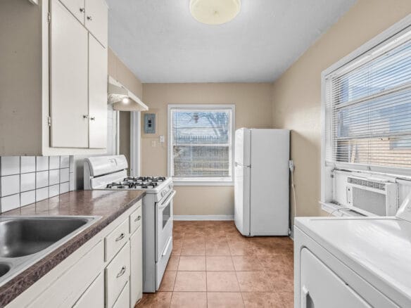 Lubbock Lease Homes: A small kitchen with white cabinets, a gas stove, double sink, white refrigerator, window with blinds, beige walls, tiled floor, and a washing machine next to a window air conditioner.